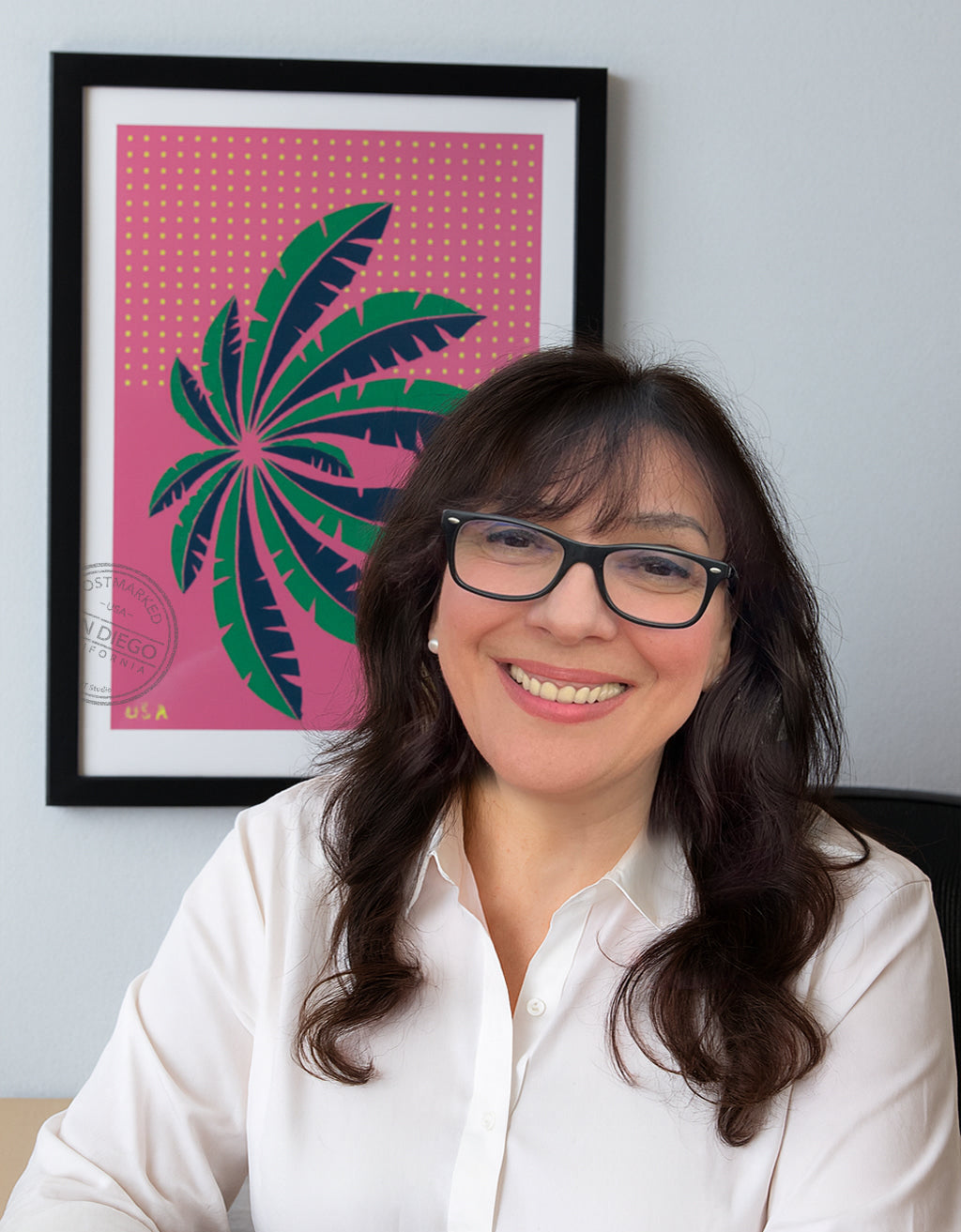 Ana is wearing glasses and a white shirt, sitting in front of a framed artwork of a stamp with a palm tree design.