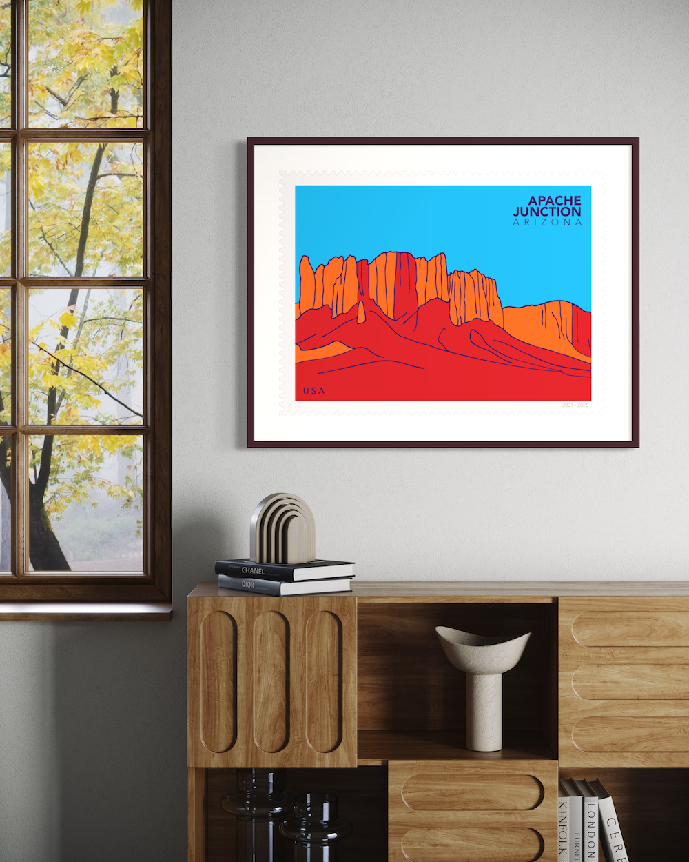 Large postage stamp with stylized illustration of desert cliffs with 'Apache Junction, Arizona' text on a blue background framed on the wall above the cabinet, by the window.