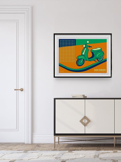 Framed artwork of a scooter stamp on an orange background above a white cabinet.