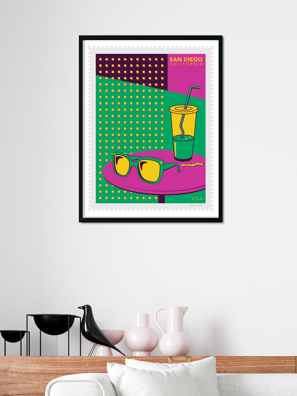 Framed colorful poster with sunglasses and a drink on a white wall above a wooden shelf.