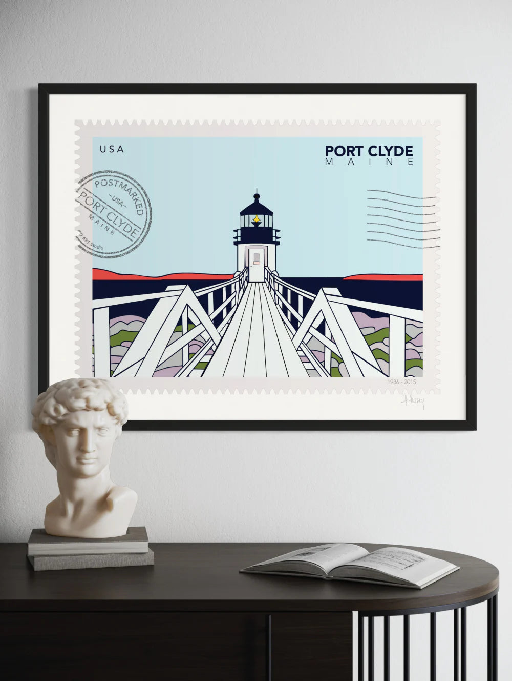 Drawing of Marshall Point Lighthouse in Port Clyde, Maine, in the winter. The drawing looks like a postage stamp with a postmark and words Port Clyde, Maine. Framed on a wall. Ana Hussey art.