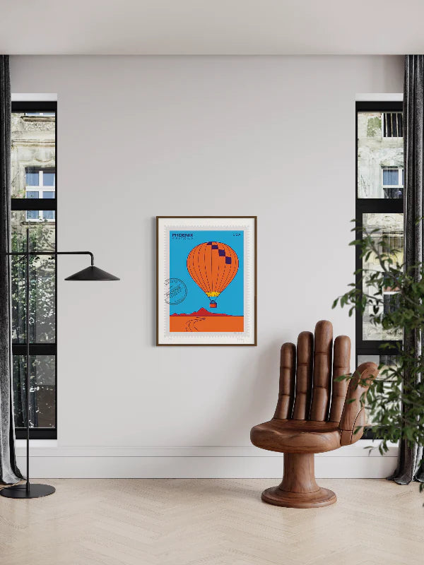 Bright colors drawing of a Phoenix hot-air-balloon ride, floating above the Phoenix desert in this large postage-stamp drawing. Drawing includes words Phoenix Arizona and postmark. Framed on a wall between two windows and a chair shaped like a hand. Ana Hussey art.