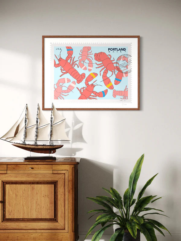 Drawing of lobsters on a blue background. The drawing looks like a postage stamp with a postmark and words Portland, Maine. Framed on a wall above a wooden cabinet with a ship model and a plant. Ana Hussey art. 