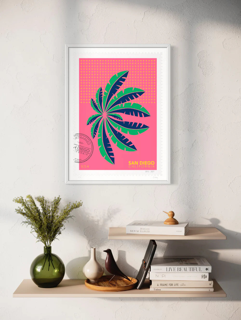 A drawing of a green San Diego palm in Mission Bay swaying against a bright pink sky. The drawing is like a large postage-stamp with postmark and includes words San Diego, California. Framed on a wall. Ana Hussey art.