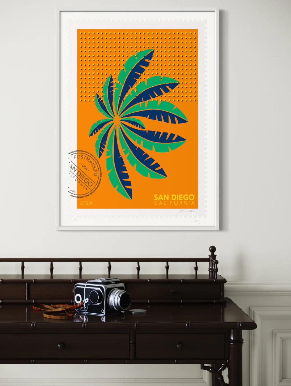 A drawing of a green and blue San Diego palm in Mission Bay swaying against a bright orange sky. The drawing is like a large postage-stamp with postmark and includes words San Diego, California. Framed on a wall. Ana Hussey art.