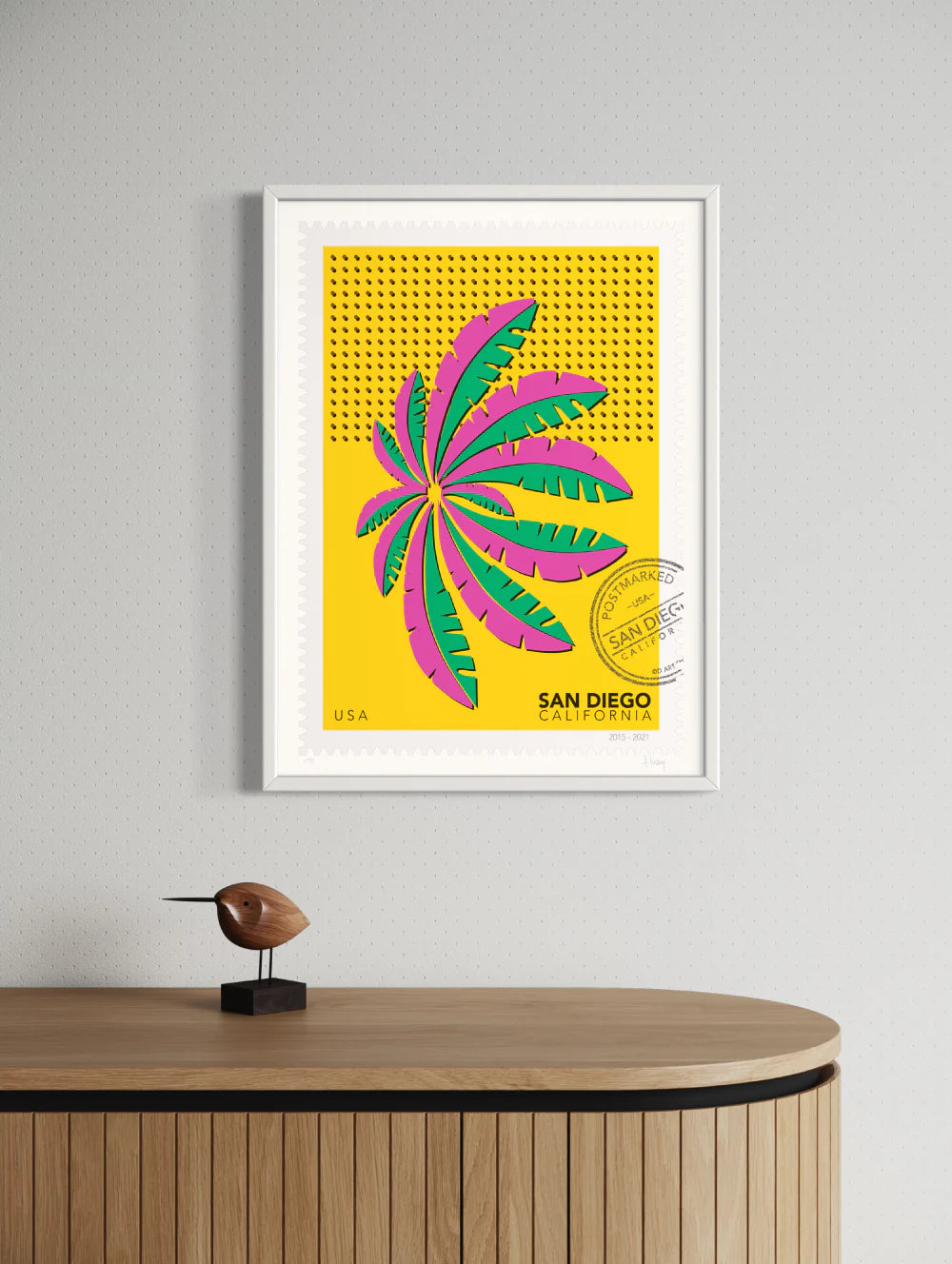 A drawing of a green and blue San Diego palm in Mission Bay swaying against a bright yellow sky. The drawing is like a large postage stamp with a postmark and includes words San Diego, California. Framed on a wall. Ana Hussey art.