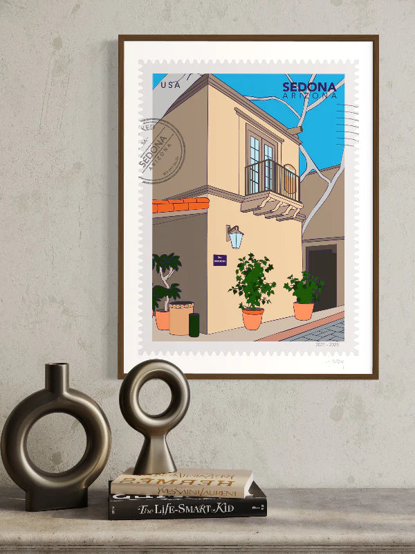 A large postage-stamp style drawing of Sedona showing the charming streets of Tlaquepaque Art Village. Sunlight spills across sandstone arches and terracotta walls, potted flowers in the corners. Includes words Sedona, Arizona and it is postmarked. Framed on a wall. Ana Hussey art.