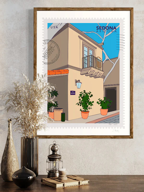 A large postage-stamp style drawing of Sedona showing the charming streets of Tlaquepaque Art Village. Sunlight spills across sandstone arches and terracotta walls, potted flowers in the corners. Includes words Sedona, Arizona and it is postmarked. Framed on a wall. Ana Hussey art.