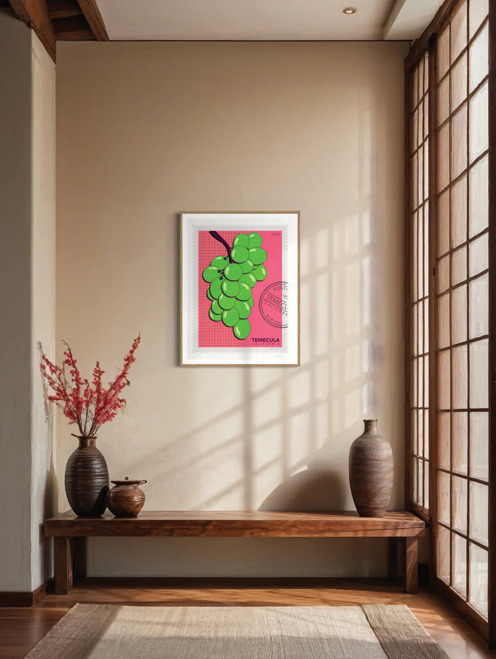 A drawing of green Temecula grapes against a bright pink background. The drawing is like a large postage stamp with a postmark and includes words Temecula, California. Framed on a wall. Ana Hussey art.