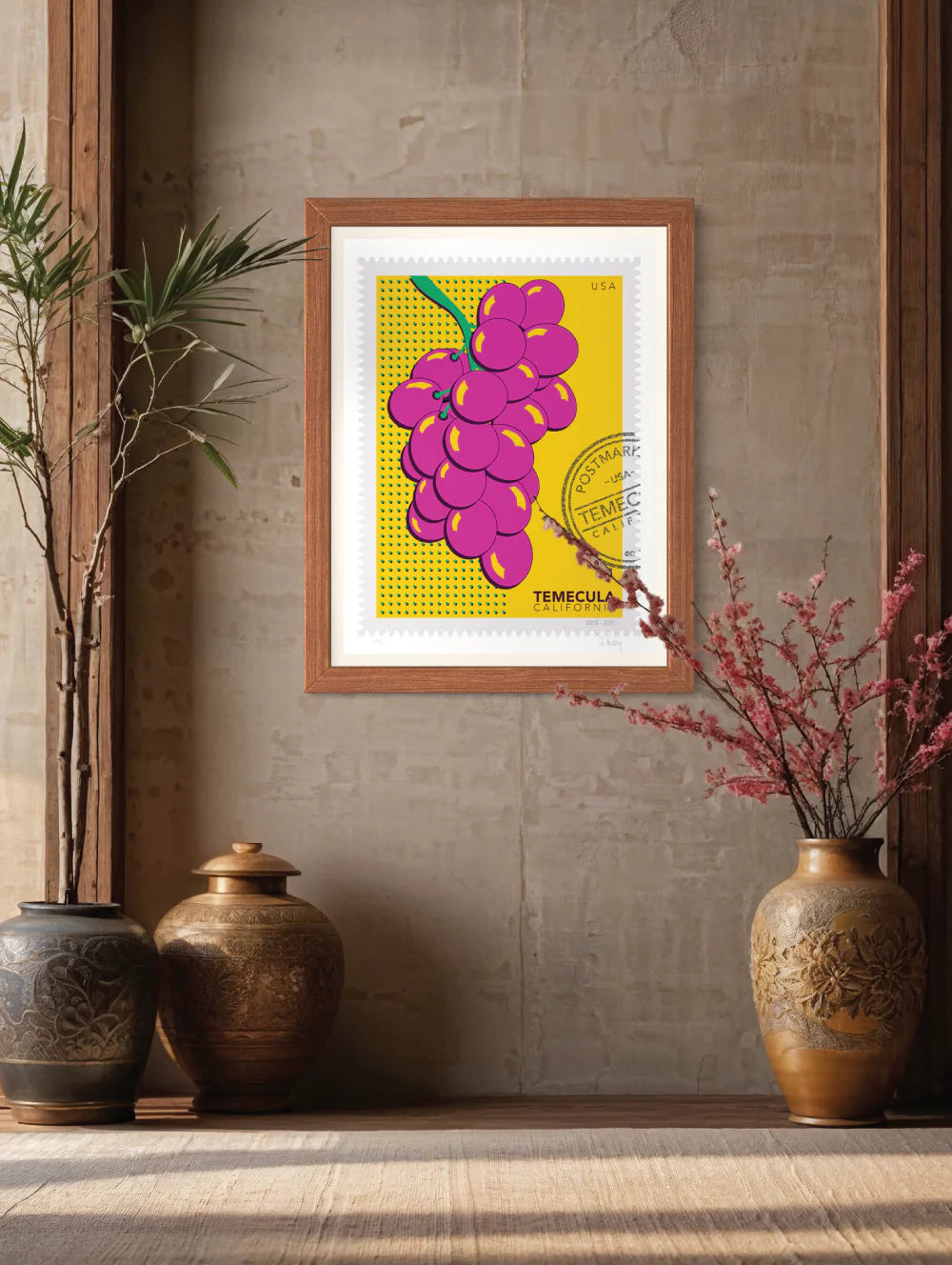 A drawing of pink Temecula grapes against a bright yellow background. The drawing is like a large postage stamp with a postmark that includes the words Temecula, California. Framed on a wall. Ana Hussey art.