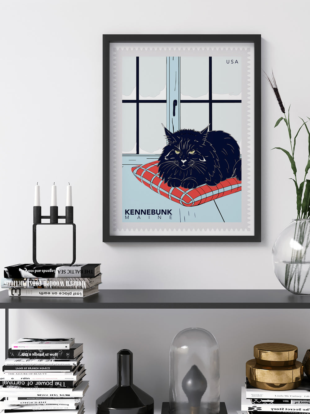 Framed artwork: postage stamp of a black cat on a red cushion with 'Kennebunk, Maine' text, displayed on a shelf.